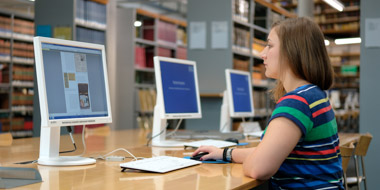 Eine Benutzerin liest eine Netzpublikation an einem unserer Leseesaal-PCs. Im Hintergrund Bibliotheksregale mit Büchern.