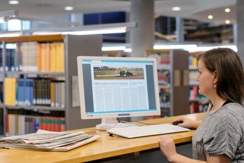 Im Lesesaal der Deutschen Nationalbibliothek liest eine Benutzerin auf dem Computerbildschirm die E-Paper-Ausgabe einer Tageszeitung. Auf dem Tisch liegen einige gedruckte Zeitungsausgaben.