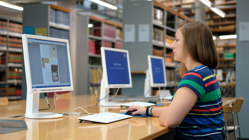 Eine Benutzerin liest eine Netzpublikation an einem unserer Leseesaal-PCs. Im Hintergrund Bibliotheksregale mit Büchern