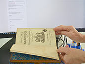 Foto eines mit Händen aufgeschlagenen Buches, im Hintergrund ist ein Bildschirm zu sehen